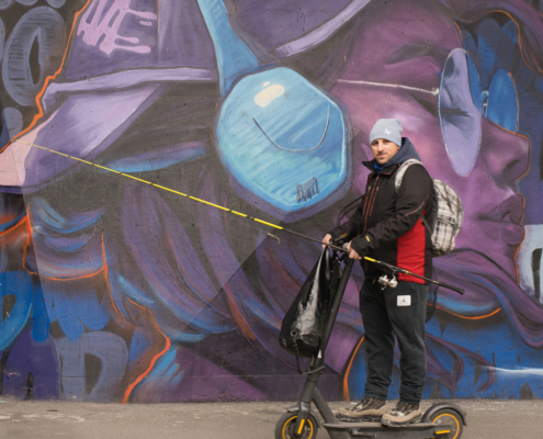 Mann auf E-Scooter mit Angel mit Blick in die Kamera vor Grafiti